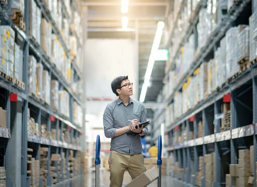 Warehousing and Logistics Insurance - Young Warehouse and Logistics Worker Completing Inventory and Stock of Product on Shelves in a Large Warehouse a Using Digital Tablet and Pen